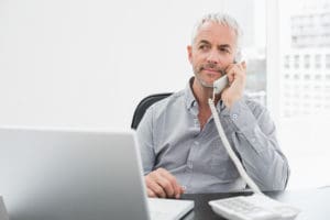 Serious mature businessman on call in front of laptop at desk in a bright office
